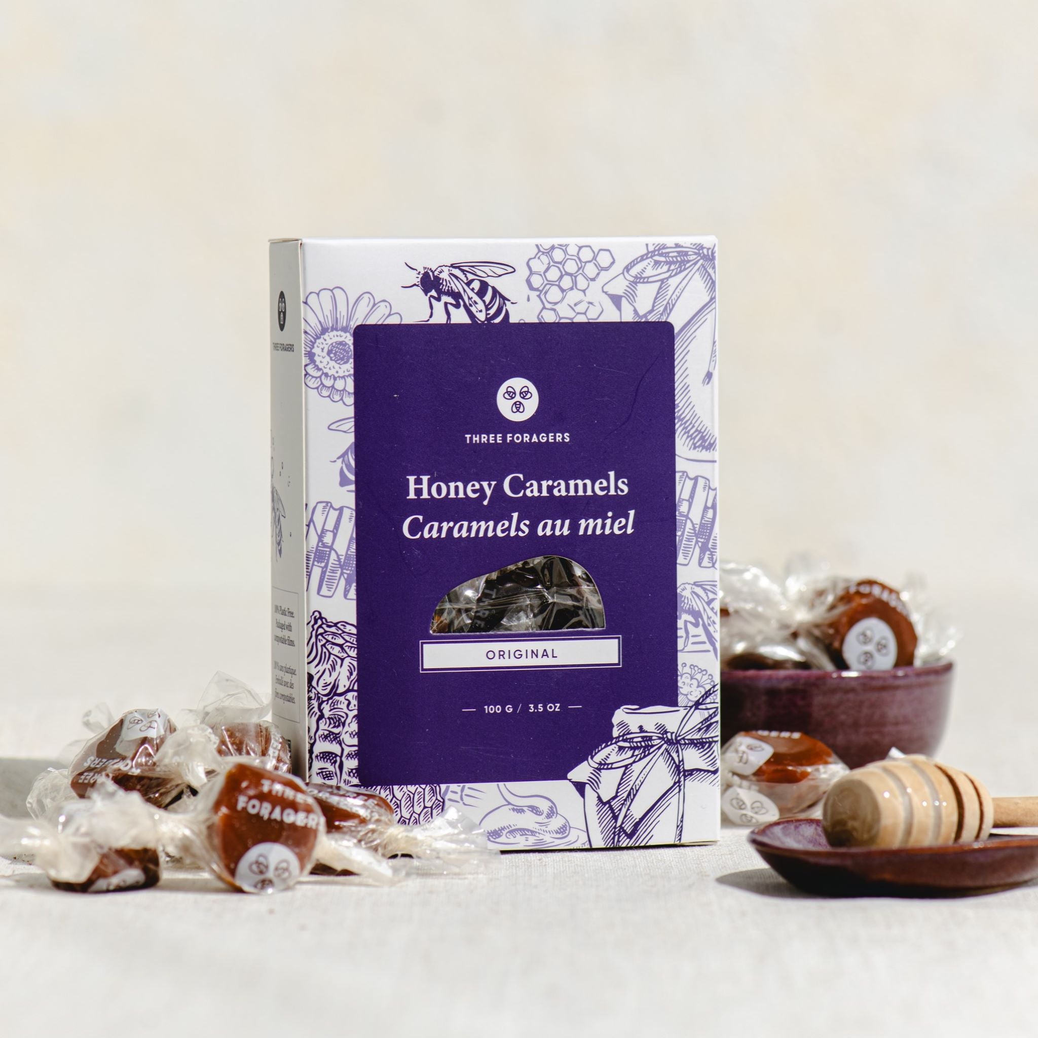 A box of Three Foragers Original Honey Caramels with some scattered wrapped honey caramels around it next to a honey dipper