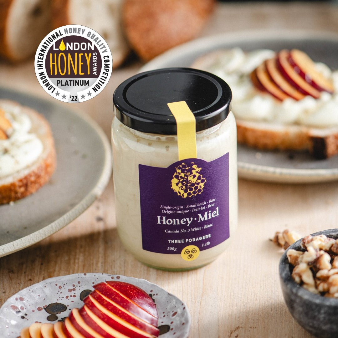 A jar of Three Foragers brand honey amid multiple plates of food containing apple slices, walnuts, and toast
