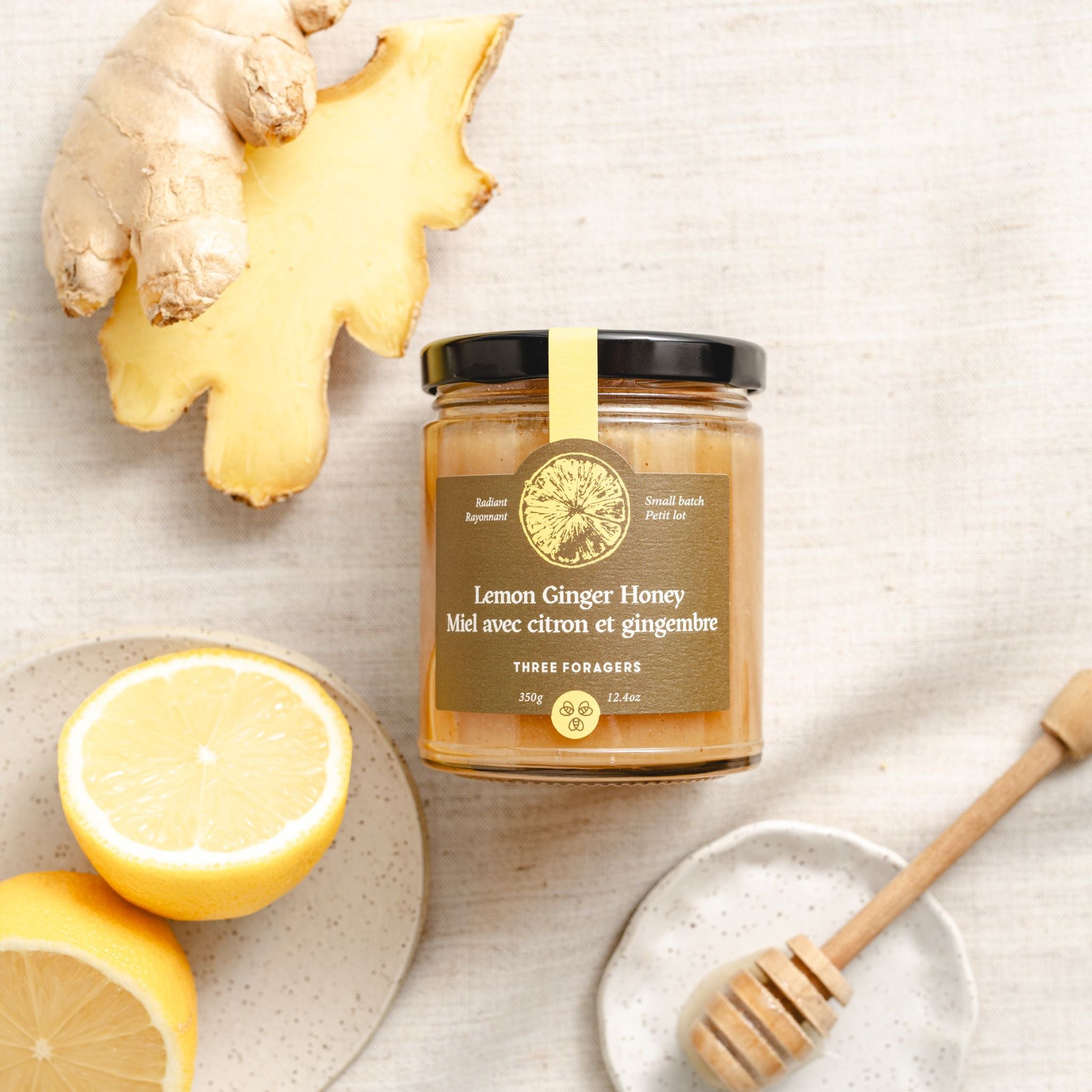A jar of Three Foragers Lemon Ginger Honey between a slice of lemon and a piece of ginger root with a honey dipper off to the side