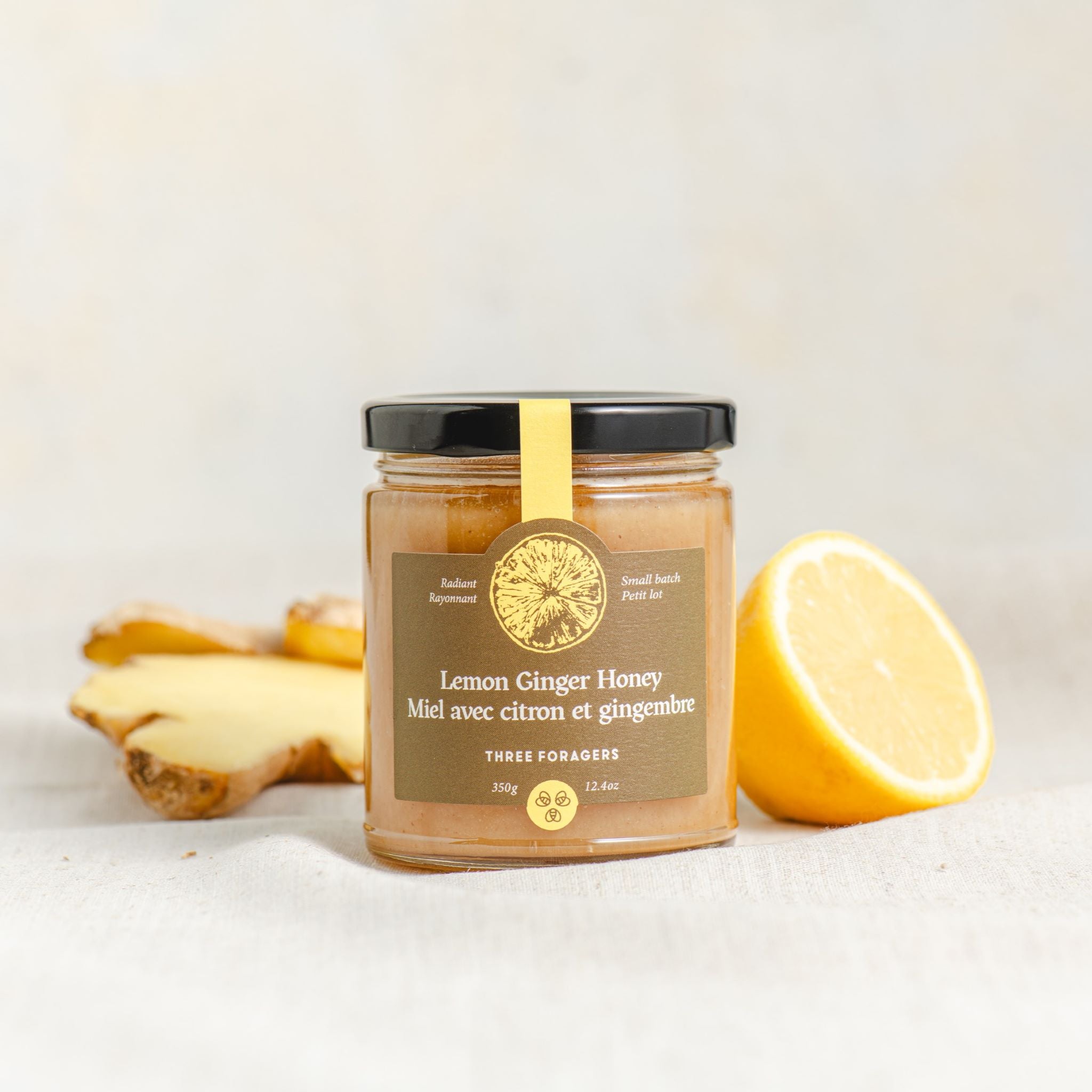A jar of Three Foragers Lemon Ginger Honey between a slice of lemon and a piece of ginger root