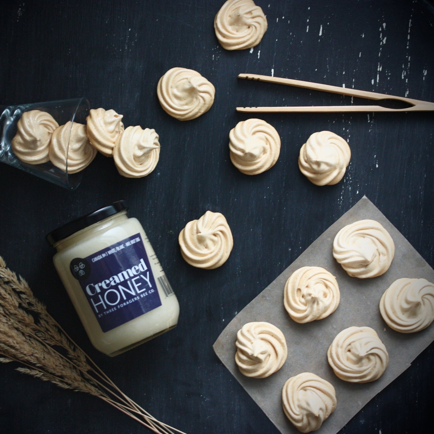 A black tabletop with a jar of Creamed Honey surrounded by a few stalks of wheat, some Honey Vanilla Bean Meringues, and a set of wooden tongs.