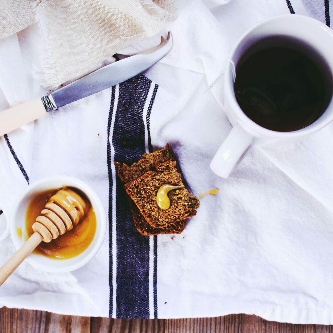 A cloth spread with a piece of bread with a drip of honey on it, a honey dipper in a dish of honey, and a cup of tea