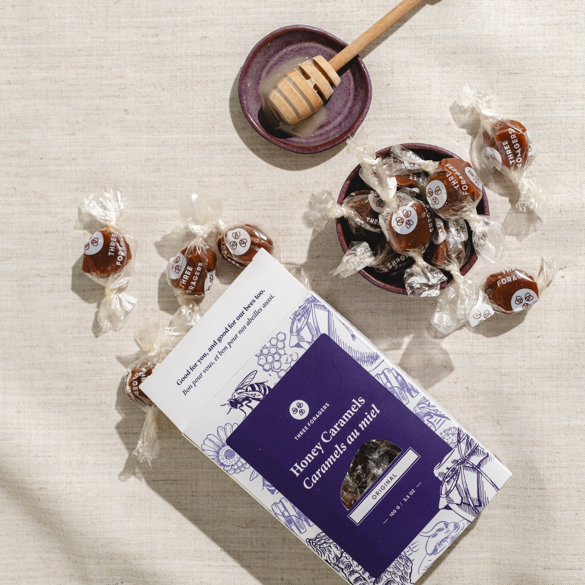 A dish of wrapped honey caramels next to a box of Three Foragers Original Honey Caramels and a honey dipper