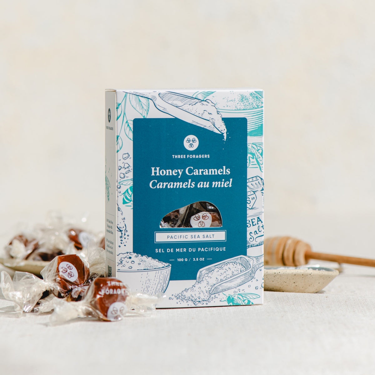 A box of sea salt honey caramels with some wrapped honey caramels and a honey dipper around it